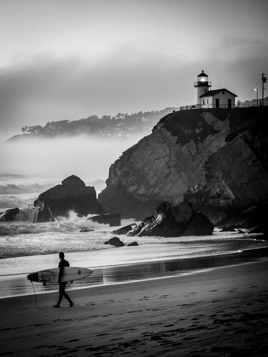 Surfer walking the beach below the Santa Cruz lighthouse on a foggy evening